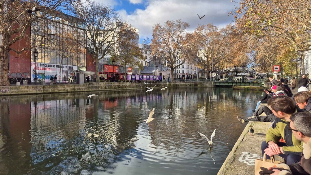 A calm river with swans and seagulls, people sitting on the riverbank, and buildings in the background. 5