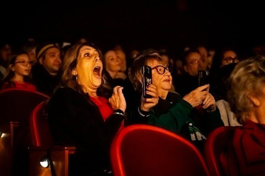 People seated in a theater with red chairs, some holding up their phones. 2
