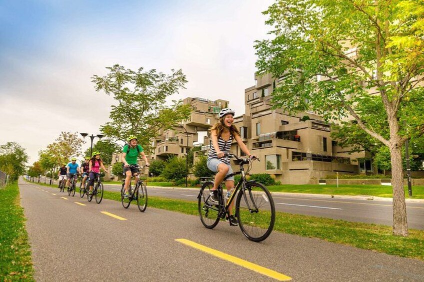 A group of cyclists on a paved path with trees and modern buildings in the background. 1