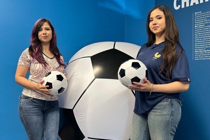 Two individuals holding soccer balls in front of a large soccer ball and a wall with text. 3