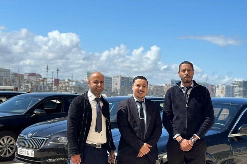 Three men standing in front of a black car with a cityscape in the background. 4