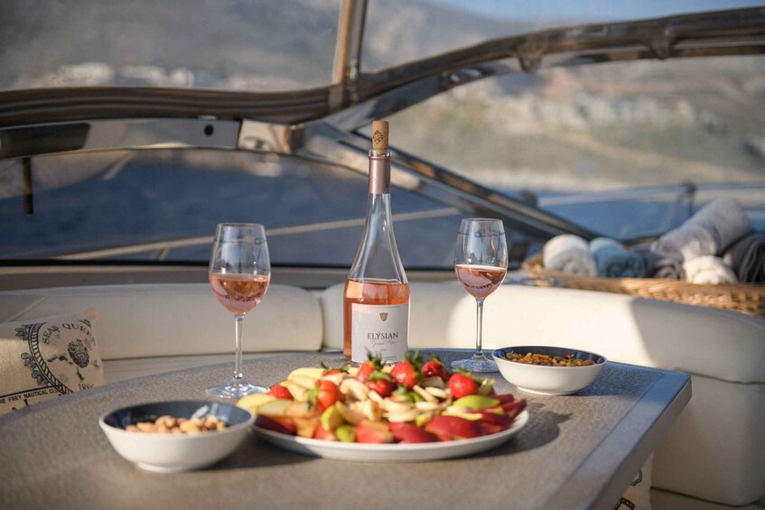 A table with a bottle of Elysian wine, two glasses, a fruit platter, and a bowl of nuts on a boat deck. 1