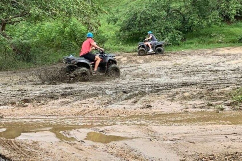 Two people riding ATVs on a muddy trail in a forested area. 4