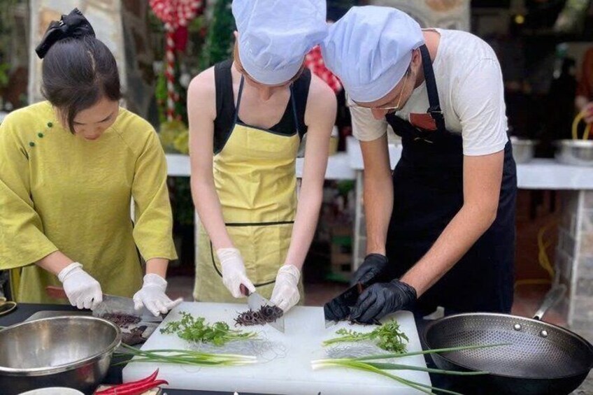 Three people preparing food, wearing aprons and gloves, with vegetables and a pan on the table. 4