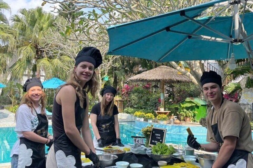 A group of people preparing food outdoors by a pool with tropical plants and a thatched-roof structure in the background. 2