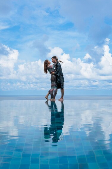 A couple embracing on a clear blue sky with clouds, standing on a reflective pool edge. 1