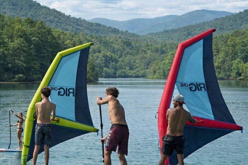 Three people with windsurfing equipment on a lake with forested hills in the background. 1