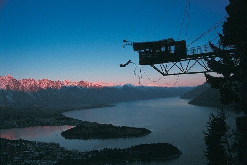 A suspension bridge with a person bungee jumping, a calm lake, and snow-capped mountains in the background. 4
