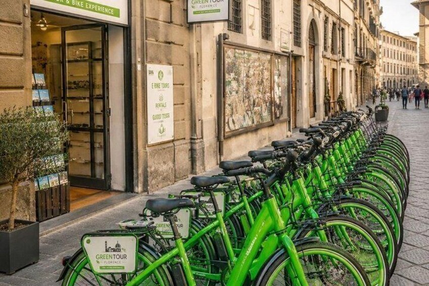 A row of green bicycles parked along a cobblestone street with a shop entrance and signage for 'GREEN TOUR.' 1