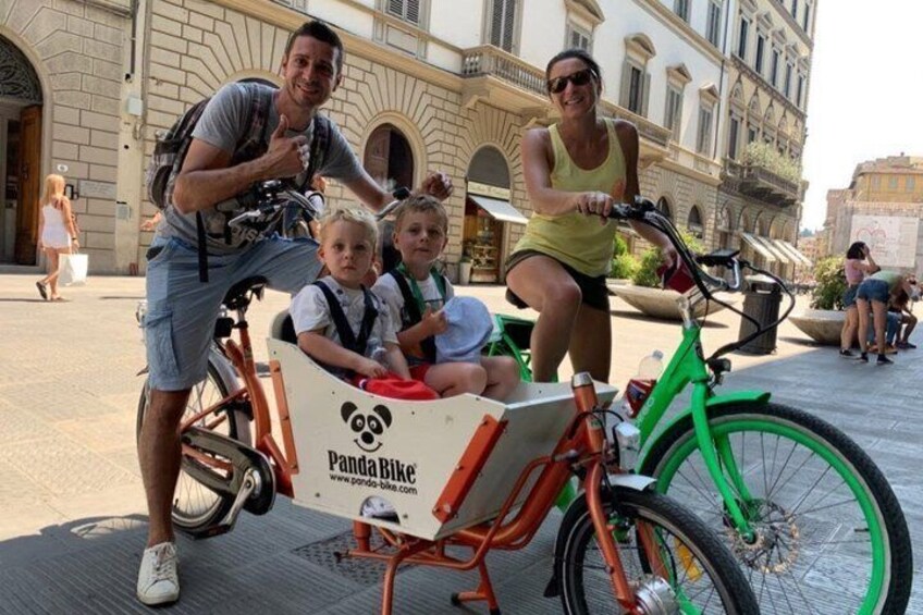 A family with a child in a Panda Bike basket, a man on a bicycle, and a woman on another bicycle in an outdoor urban setting. 4