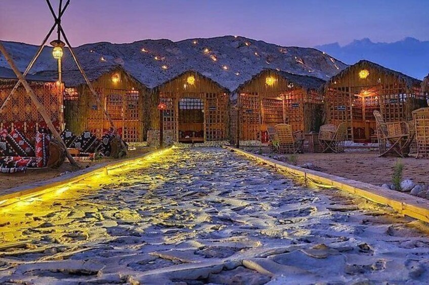 A snow-covered pathway leading to a row of wooden buildings with thatched roofs, illuminated by warm lights. 3