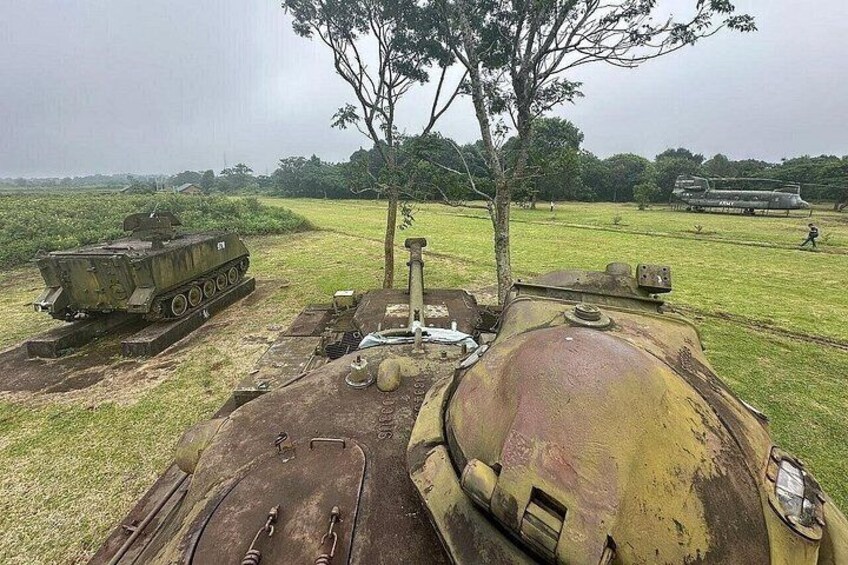 A military tank on a grassy field with a tree and another tank in the background. 4