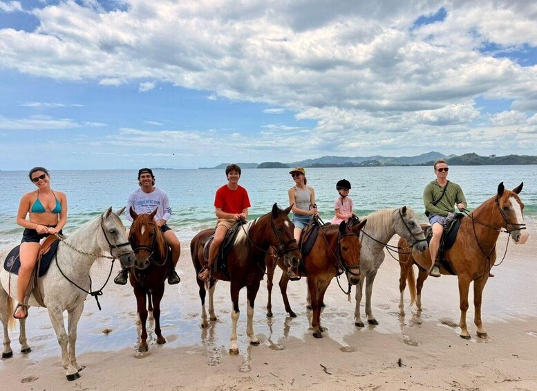 A group of people riding horses on a beach with a body of water and mountains in the background. 2