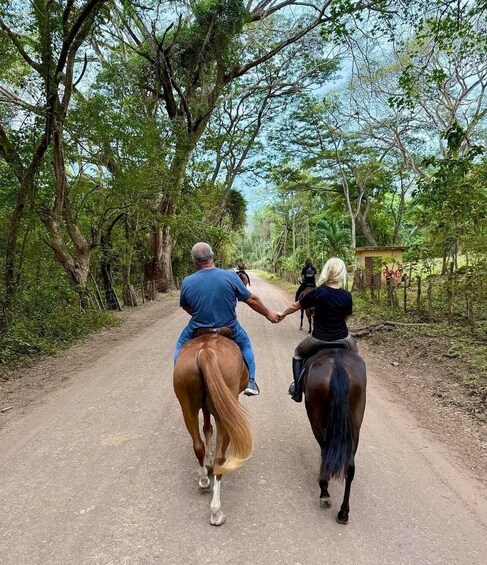 Two people horseback riding on a dirt path surrounded by trees. 4