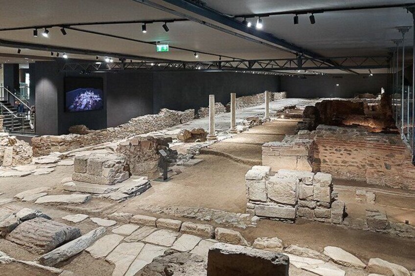 An indoor archaeological exhibit with stone ruins and a large screen displaying a cityscape. 2