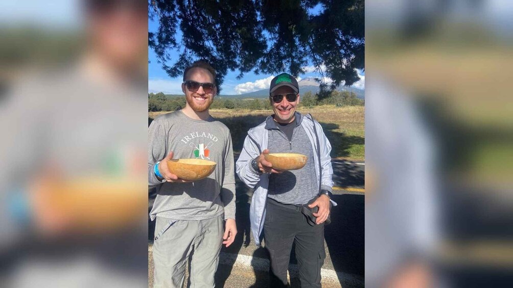Two people holding bowls outdoors with trees and a clear sky in the background. 4