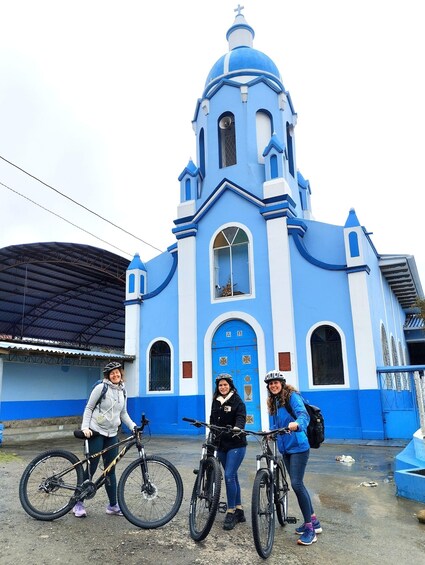 A blue church with a bell tower, a covered structure, and three people with bicycles. 3