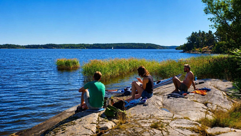 Three people sitting on a rocky shore by a lake with a clear sky. 4