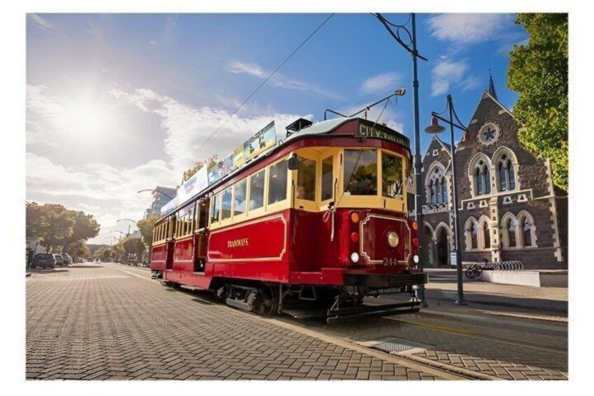 A red tram with 'CityRail' on the front, traveling on a track with a brick-paved road, passing by a historic building with arched windows. 1