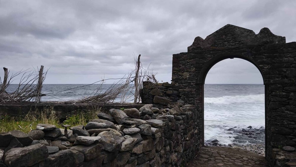 A coastal stone wall with an archway leading to the sea, surrounded by overgrown vegetation and a stormy sky. 1