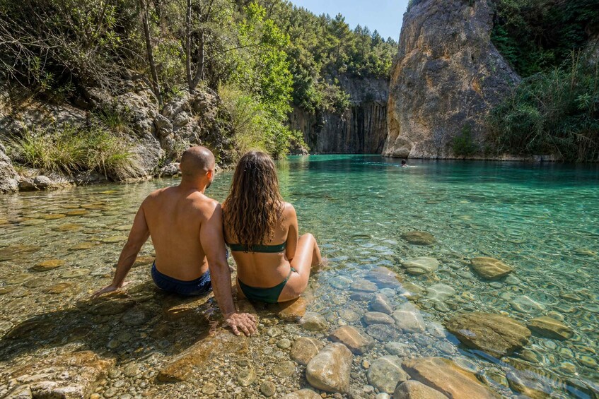 Two people sitting by a clear river surrounded by rocky terrain and greenery. 1