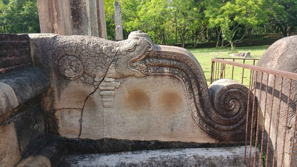 A stone structure with intricate carvings and a spiral design, surrounded by a park with trees and a metal railing. 2