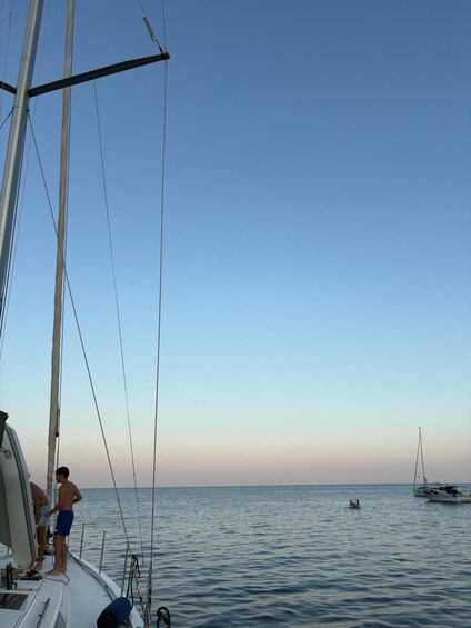 A sailboat with two people on deck, calm sea, and another boat in the distance. 2
