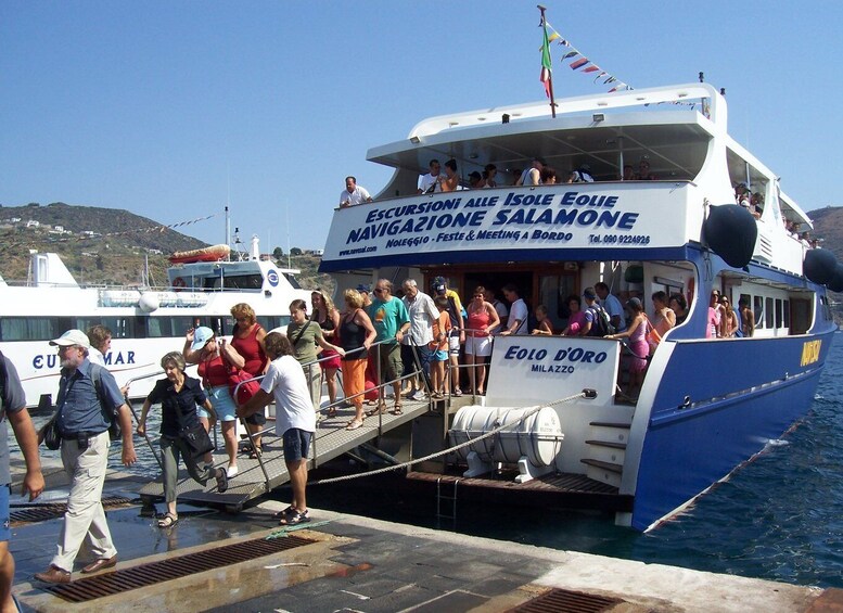 A ferry with passengers boarding, named 'ESCURSIONI ALLE Isole EOLIE NAVIGAZIO - FESTE & MEETING A BORDO.' 1