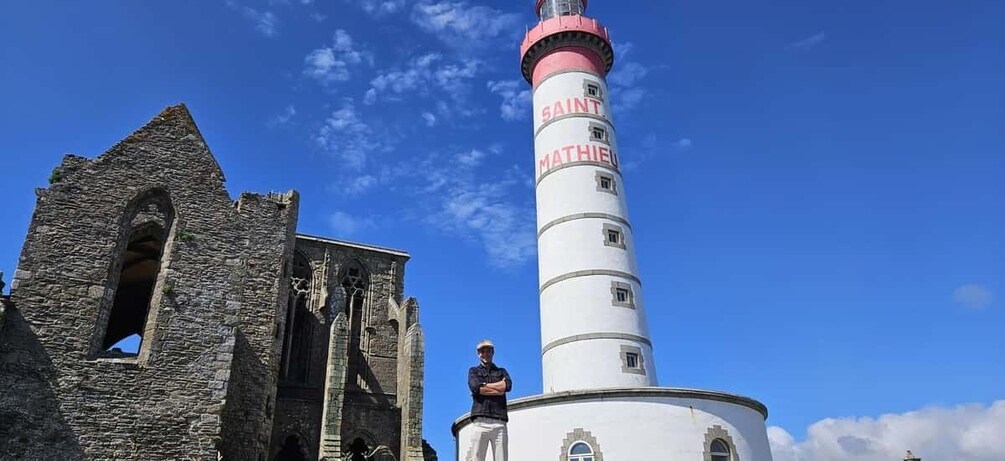 A lighthouse with 'SAINT MATHIEU' written on it, a stone building with an arched window, and a clear blue sky. 1