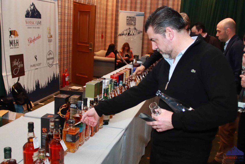 A man pouring a drink from a bottle at a bar with various liquor bottles and branded boxes. 2