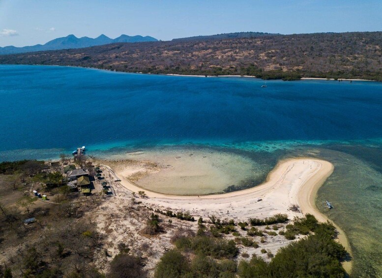 An aerial view of a coastal area with a curved sandy beach, clear blue waters, and a mountainous backdrop. 1