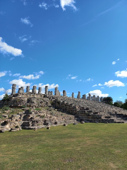 Ancient stone ruins with stepped pyramids on a grassy hill under a clear blue sky. 1