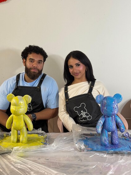 Two people wearing aprons with bear designs, holding bear-shaped objects, and plastic bags on a table. 1