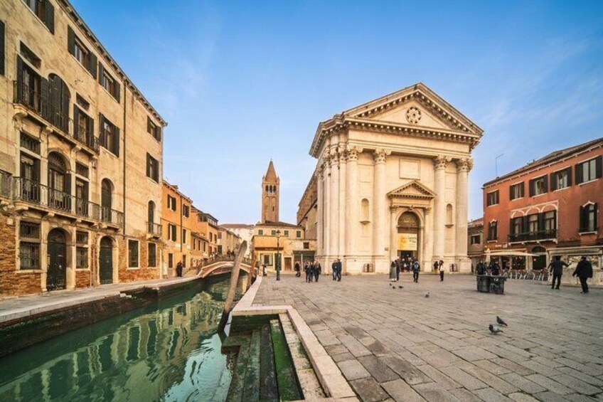 A canal with buildings on either side, a prominent church with a clock tower, and people walking in the square. 5