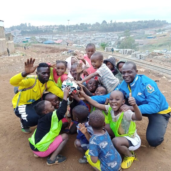 A group of children and adults gathered on a dirt ground with a railway track and industrial buildings in the background. 3