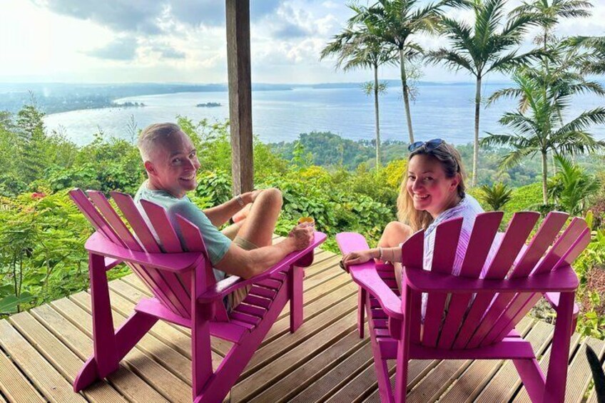 Two people sitting on pink Adirondack chairs overlooking a scenic view of a body of water and palm trees. 2