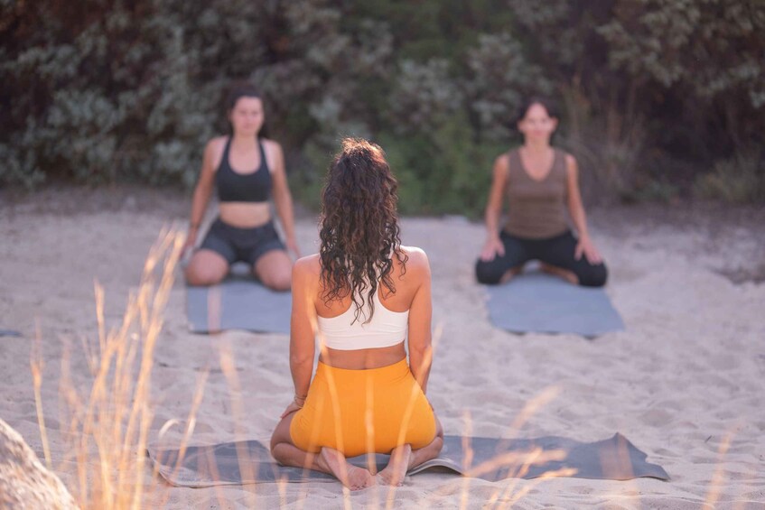 Three individuals sitting on yoga mats on a sandy beach with vegetation in the background. 1