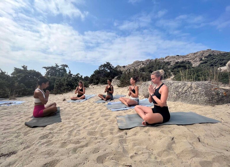Four people meditating on yoga mats on a sandy beach with a rocky landscape in the background. 2