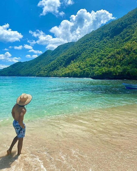 A person standing on a beach with clear turquoise water, a boat on the right, and a lush green mountain in the background. 3