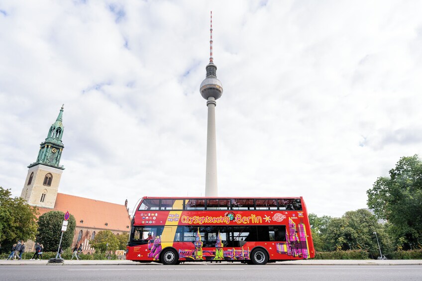 Punainen kaksikerroksinen bussi, jonka kylkeen on maalattu teksti ”CitySightseeing Berlin”, on pysäköity Fernsehturm-tornin ja vihreän tornin huipun omaavan kirkon eteen. 5