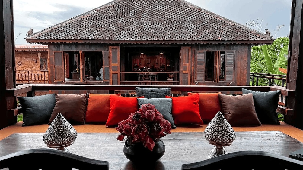 A wooden house with a tiled roof, a balcony with a sofa and cushions, and a table with a vase of flowers. 1