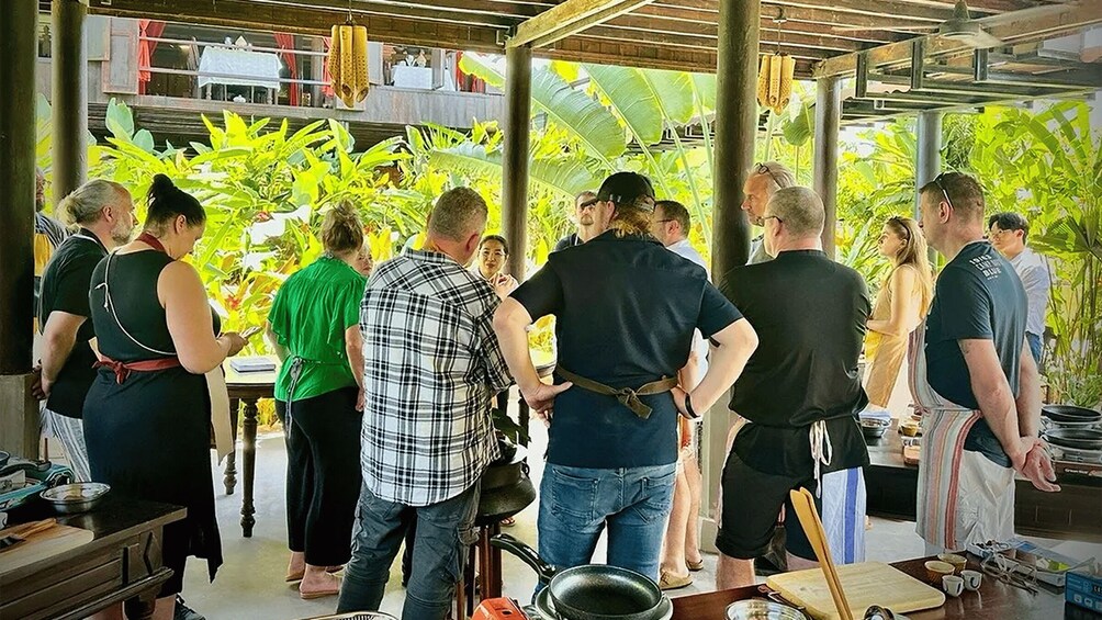 A group of people in a covered outdoor area with tables and cooking equipment, surrounded by lush greenery. 4