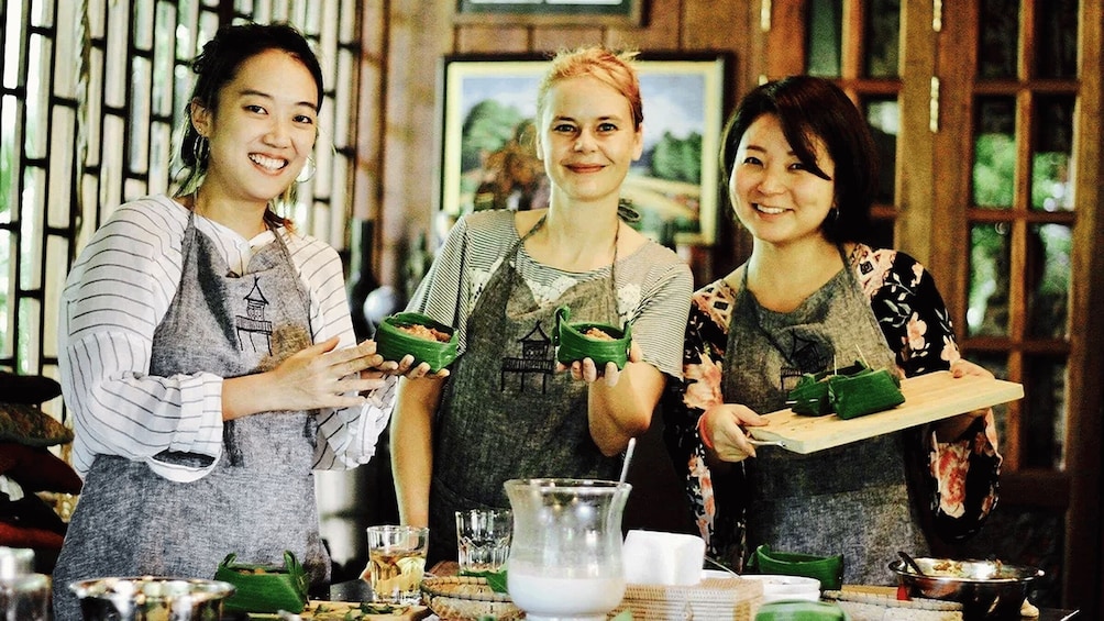 Three people in a kitchen holding green containers, with cooking utensils and a glass pitcher on the counter. 3