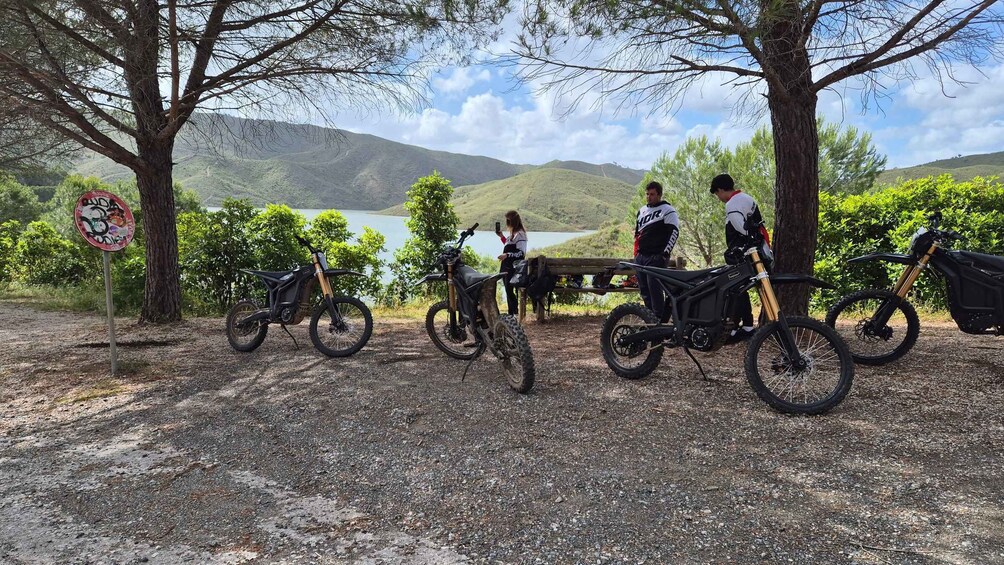 A gravel path with three dirt bikes parked, a lake, and hills in the background. 5
