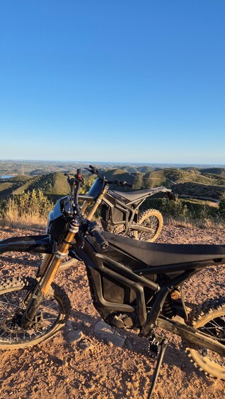 Two dirt bikes parked on a rocky terrain with a scenic view of hills and a clear blue sky in the background. 4