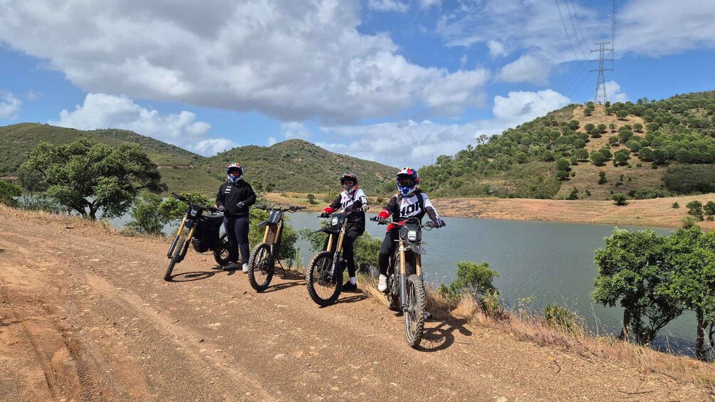 Three mountain bikers on a dirt path beside a lake with hills in the background. 1