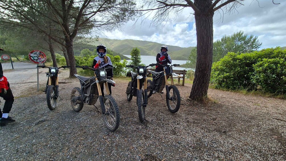 Three dirt bikes parked on a gravel path with a lake and hills in the background. 2