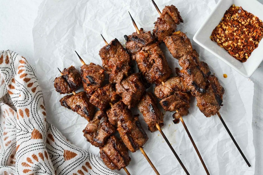 Skewered meat on a white surface with a patterned cloth and a bowl of chili flakes. 5