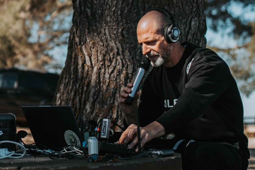 A person sitting outdoors with a laptop and audio equipment, surrounded by cables and a microphone. 5