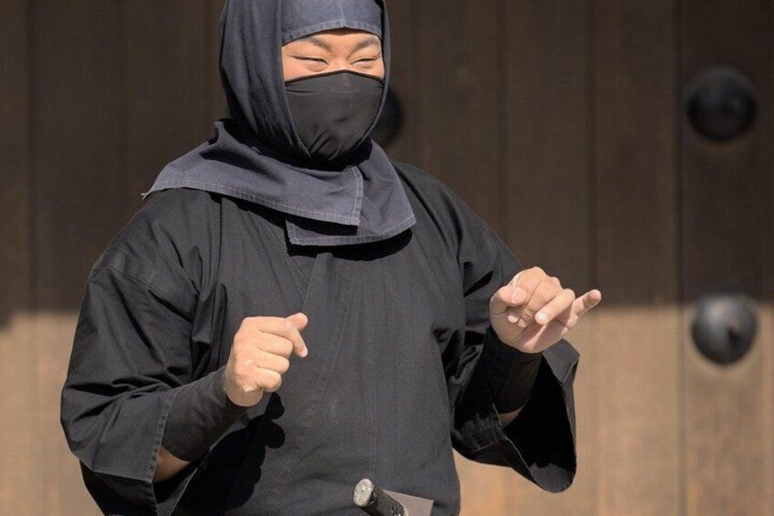 A person in a black outfit with a hood, pointing with both hands. 1